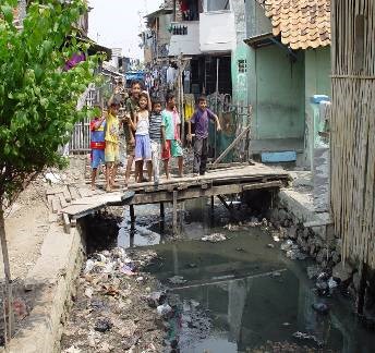 Children on a wooden bridge over a dirty drain in a crowded urban slum area.